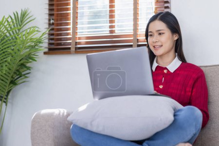Happy casual beautiful Asian woman is talking on a laptop computer sitting on a sofa at home. mug #625552666