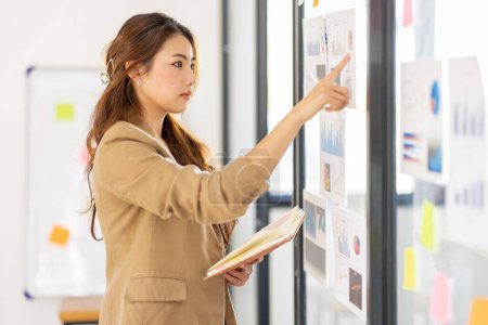 Téléchargez les photos : Young Business Asian woman using sticky notes and graph chart in glass wall analyzing strategy business plan to development grow to success - en image libre de droit