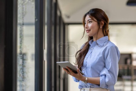 Téléchargez les photos : Young business Asian woman using tablet and standing near the window in workplace - en image libre de droit
