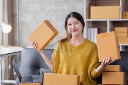 Foto de Startup SME small business entrepreneur of freelance Asian woman using a laptop with box Cheerful success Asian woman her hand lifts up online marketing packaging box and delivery SME idea concept - Imagen libre de derechos
