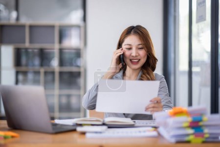 Téléchargez les photos : Smiling young Asian woman sitting and call phone device, looking at laptop, checking modern apps, texting messages, browsing internet doing shopping relaxing at home. - en image libre de droit