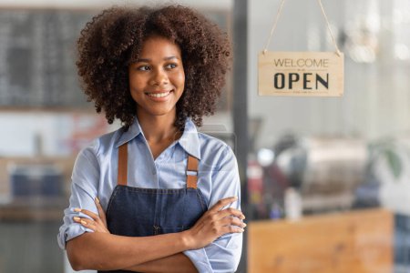 Téléchargez les photos : Happy waitress standing at restaurant entrance. Portrait of african american business woman attend new customers in her coffee shop. Happy woman owner showing open sign in her small business shop. - en image libre de droit