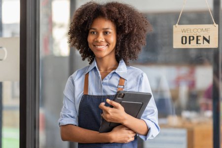 Photo for Successful african woman in apron standing coffee shop door. Happy small business owner holding tablet and working. Smiling portrait of SME entrepreneur seller business standing with copy space. - Royalty Free Image