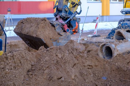 Téléchargez les photos : Une excavatrice travaille sur un chantier de construction pour remplacer un pipeline en hiver. Creuser des trous pour la pose de nouveaux tuyaux pour le chauffage central dans un quartier résidentiel. Un tuyau en béton porté dans le sable. - en image libre de droit