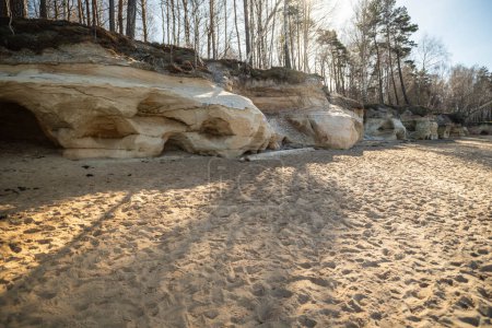 Eine ruhige Landschaft mit erodierten Felsen inmitten eines sandigen Geländes und der Kulisse eines sonnenbeschienenen Waldes. Klippen von Veczemju, Lettland