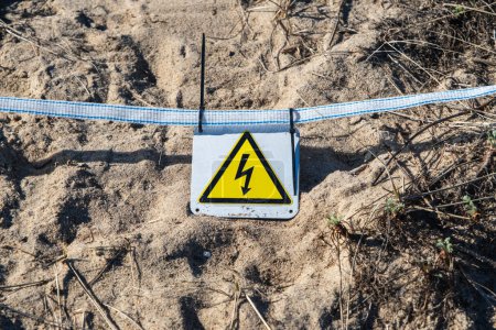 A yellow triangular warning sign for electrical hazard on sandy ground.