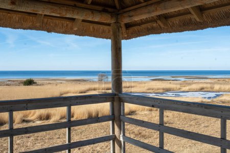 A serene view of the sea and grassland from a wooden gazebo under a clear blue sky.