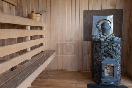Interior de una sauna tradicional de madera con un banco, un cubo y una estufa con rocas para generar calor de vapor.