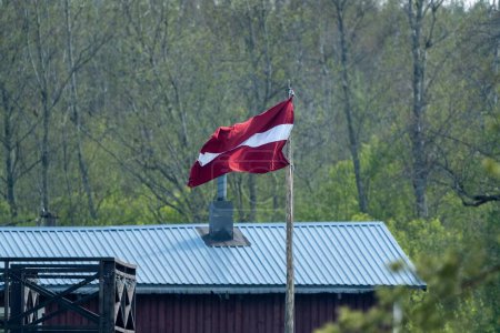 The Latvian flag waves on a wooden pole in a rural area, set against a backdrop of trees and a building with a blue metal roof.