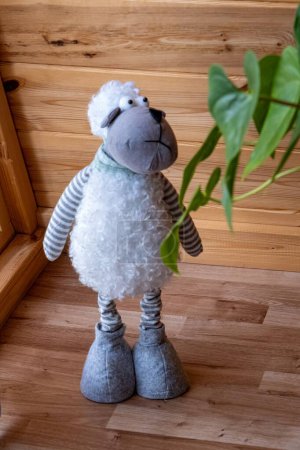 A plush toy sheep with striped legs and a curly body stands on a wooden floor in a cozy, wooden interior, next to a green plant.
