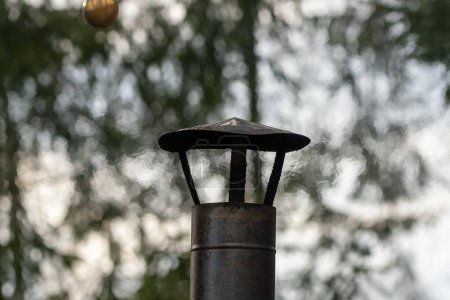 A close-up view of a rustic metal chimney cap against a blurred background of trees, highlighting an outdoor forest setting and old craftsmanship.
