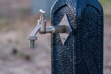 Close-up of a metal water tap mounted on a textured black stand, highlighting its outdoor placement and utility.