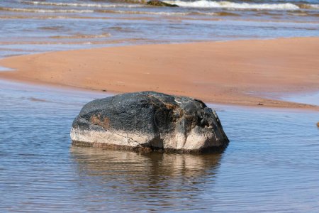 Une grande roche partiellement submergée dans les eaux peu profondes près d'une plage de sable, avec de douces vagues en arrière-plan par une journée ensoleillée.