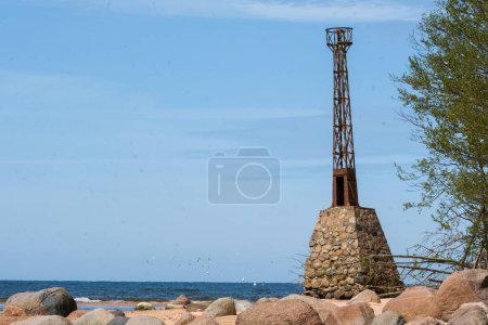 A rustic lighthouse tower stands on a rocky beach with a clear blue sky and ocean in the background, highlighting coastal scenery.