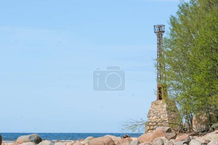 An old lighthouse tower stands beside the ocean shore, surrounded by rocks and greenery, under a clear blue sky.