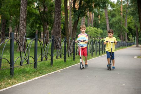 Zwei attraktive europäische Burschenbrüder in rot-weiß karierten Hemden stehen auf Motorrollern im Park. Sie lachen, lächeln, umarmen sich und haben Spaß. Aktive Freizeit mit Kindern.