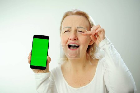 Photo for An adult woman in a white T-shirt holds a phone with a green chromakey screen in her hands, she cries with joy, wipes a tear with her finger and is very happy Advertise your content - Royalty Free Image