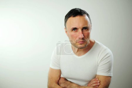 Foto de Man looks into frame he leaned forward little and crossed his arms over his chest on him white t-shirt look expresses strictness seriousness a gaze takes piercing eyes doubts eyebrows slightly frowned - Imagen libre de derechos