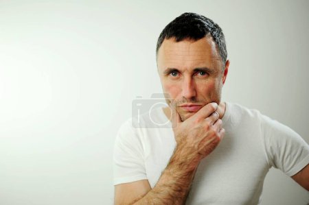 Téléchargez les photos : Adult man thought He put hand on chin and confidently looks with piercing gaze into frame he has dark hair with gray hair he is in white T-shirt on white background hairy hands unshaven on face - en image libre de droit