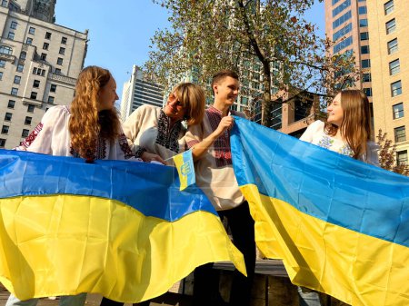 Foto de Four Ukrainian people in embroidered shirts stand on square with two flags of Ukraine happily end of war peace in Ukraine return of emigrants refugees family reunited three women one guy - Imagen libre de derechos