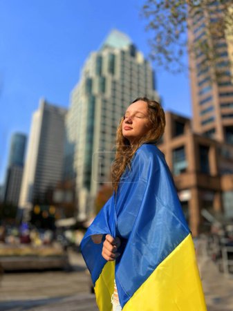 girl teenager stands wrapped in flag misses Ukraine Vancouver Canada refugees migrants war in Ukraine. Russia aggressor. Dream of end of war we For peace on shoulders flag blurred buildings of city