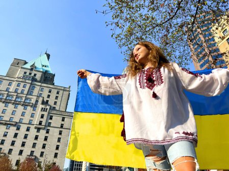 young girl teenager woman holding flag of Ukraine behind back wearing large wide white embroidered shirt with red embroidery torn blue jeans immigration to Canada war in Ukraine feeling for people
