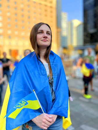 Photo for Young woman stands on square during demonstration against war she holds flag of Ukraine on shoulders flag of Ukraine emblem of trident She looks into distance war in Ukraine agressor clasps hands - Royalty Free Image