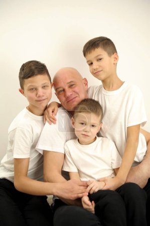 Foto de Photo of father three sons children stuck around dad from all sides it is difficult for father to keep them all in white t-shirts on white background adoption concept joy help family large family - Imagen libre de derechos