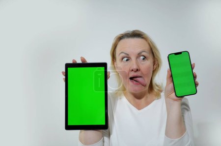Photo for Adult woman looks sideways at one of tablets hands she holds phone and tablet with green chromakey screen she is happy surprised cheerfully playfully looks away at phone space for text interest sale - Royalty Free Image