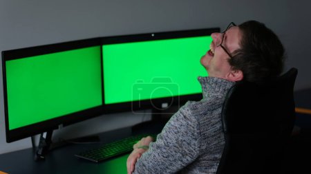 Foto de Middle-aged man in gray sweater glasses laughs very sincerely strongly he points finger throws head up sits Near table on chair in front two screens 2 monitors with green chromakey screen text ad - Imagen libre de derechos