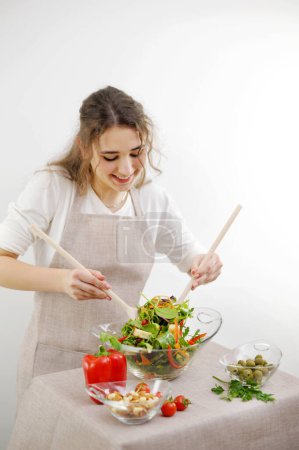 Foto de Young beautiful teenage girl stirs diligently salad leaned over bowl smiling she likes cooking healthy food vegetarian ingredients green lettuce leaves wooden spoons apron and tablecloth beige color - Imagen libre de derechos