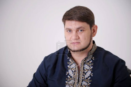 Foto de Stern look of blue eyes handsome man in Ukrainian embroidered shirt looks straight into frame he has light beard compressed lips thick hair a blue shirt and embroidery on white background close-up - Imagen libre de derechos