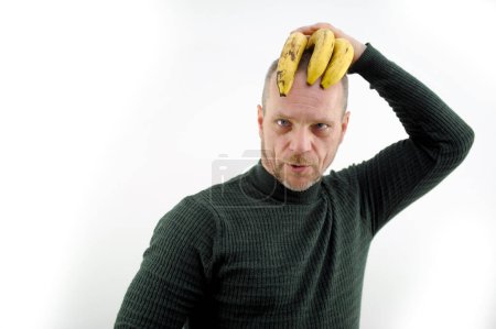Téléchargez les photos : Comic photo of man holding three spoiled bananas on his head as a hairstyle squinted eyes squint redhead with a gray beard and mustache green sweater white background make faces make faces - en image libre de droit