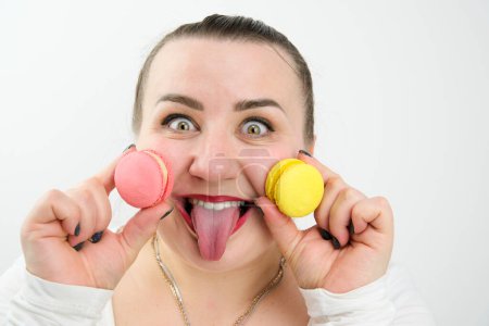 Photo for Plump cheerful woman stuck out her tongue holds two macarons in hands bake eyes laugh red lipstick white clothes hair tied up funny photo food sweets calories on a white background - Royalty Free Image