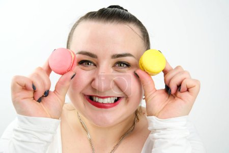 Téléchargez les photos : Une femme insatiable graisse sourit heureux tenant deux macarons de couleurs différentes dans les mains mettre le dessert sur son visage, la nourriture est douce et nocive - en image libre de droit
