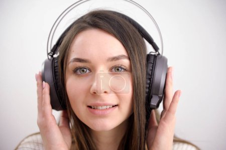 Photo for Brunette young woman listening her favourite songs in headphones looking funny on isolated white background. The girl in headphones High quality photo - Royalty Free Image