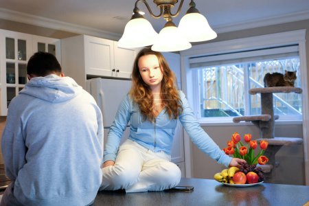 Photo for Teens sit on table in kitchen girl offended upset pouted boy turned back on phone looks next gadget eat fruit year in background near window adolescence quarrel of discontent offended at each other - Royalty Free Image