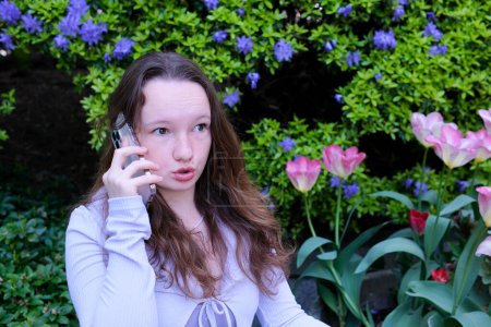 Téléchargez les photos : Indignation et mécontentement adolescent fille parler au téléphone dans la rue au printemps tulipes fleurs parler jurant yeux grands ouverts jeune femme Que s'est-il passé - en image libre de droit