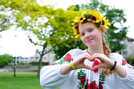 a Ukrainian woman in embroidered shirt with a sunflower wreath on her head makes a heart with her hands recognition approval joy Nadezhda pleasant emotions red-haired beauty on the street