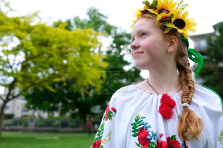 Téléchargez les photos : Jeune fille aux cheveux roux dans une couronne de tournesols à l'extérieur dans le jardin du parc en belle chemise ukrainienne brodée vyshyvanka avec tresse rouge avec rubans Ukraine Temps paisible de joie et de paix - en image libre de droit