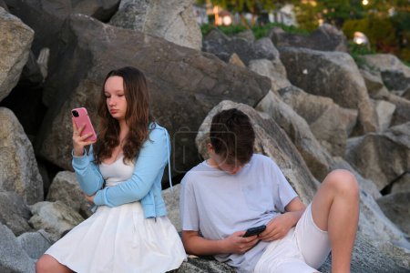 Téléchargez les photos : Adolescents assis sur des pierres un garçon et une fille avec un téléphone dans leurs mains utilisent des gadgets dans la nature dépendant des réseaux sociaux ennui sur la première date échoué communication vêtements légers adolescence - en image libre de droit