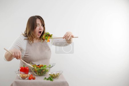 Téléchargez les photos : Funny cheerful woman eating salad on white background she opens mouth wide bulges eyes large wooden spoon stuffs portion of food on table ingredients space for text weight loss ad - en image libre de droit