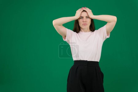 Portrait of concerned tired young woman in green casual clothes looking camera, putting hands on head isolated on bright blue wall background in studio. People lifestyle concept. 