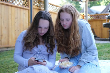Two girls friends on a picnic drinking orange juice taking a selfie talking sitting on their phones sitting on the grass in summer dresses. Two friends picnic
