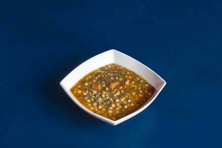 Photo for A cooked vegetarian dish of chickpeas with spinach parsley and carrots served on a deep diamond-shaped white ceramic plate on a blue studio background. - Royalty Free Image