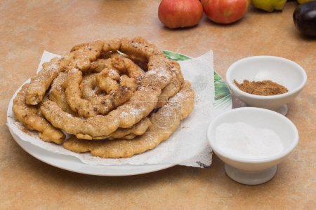 Photo for A white plate with a green leaf pattern around the edge, holding fried golden-brown freshly fried dough twists, with two small bowls of powdered sweetener and cinnamon powder on the side. - Royalty Free Image