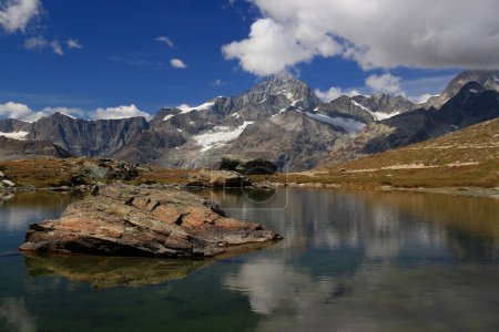 Foto de Paisaje con una superficie lisa del lago Riffelsee, montañas y nubes reflejadas en él, con una gran piedra en primer plano, en una montaña Gornergrat, cerca de Zermatt, en el sur de Suiza - Imagen libre de derechos