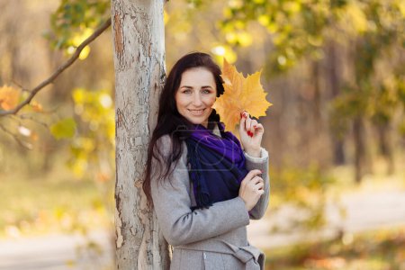 Téléchargez les photos : Une belle fille aux cheveux foncés, un manteau mouillé et une écharpe violette tient une lettre. fond flou et feuilles floues visibles ci-dessus - en image libre de droit