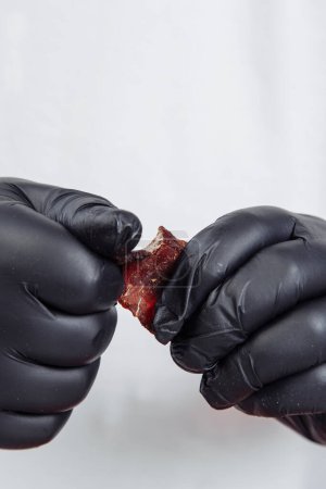 Foto de Un tipo con guantes negros rompe un pedazo de carne seca y curada sobre un fondo blanco. Foto de alta calidad - Imagen libre de derechos
