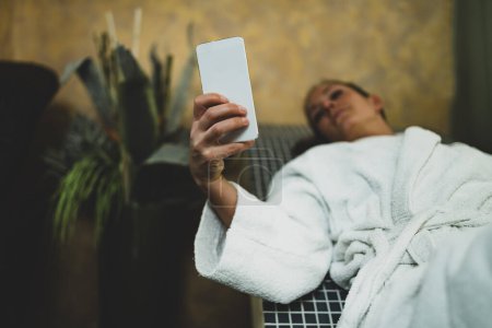 Téléchargez les photos : Femme en peignoir Allongé sur un lit de tepidarium et utilisant un téléphone intelligent
. - en image libre de droit